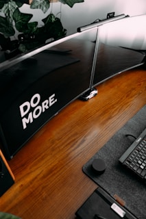 A neatly arranged wooden desk featuring a dual monitor setup, with the prominent phrase 'DO MORE' displayed. There is a sleek black keyboard, a mouse, and a small notebook with a pen on a grey mat. Green leafy plants hang above the monitors, adding a touch of nature to the workspace.