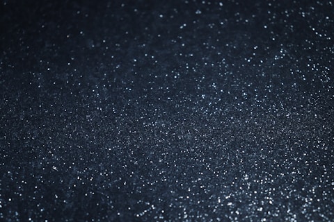 A textured surface with a shimmering effect created by numerous tiny reflective particles spread across a dark background.