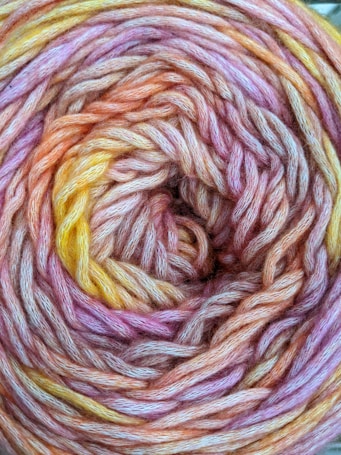 An intricate spiral pattern of wool yarn displaying a mix of vibrant colors including pink, orange, and yellow. The texture of the yarn is soft and fluffy with visible fibers intertwined closely together.