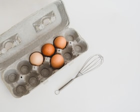 An open egg carton with four brown eggs inside is placed on a white surface. Next to the carton, there is a metal whisk.