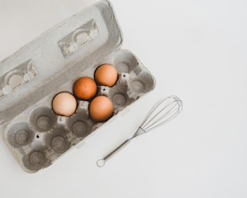 An open egg carton with four brown eggs inside is placed on a white surface. Next to the carton, there is a metal whisk.