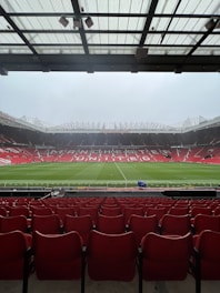A view from the stands of a large football stadium with an empty field. Rows of red seats extend towards the pitch, which is lush and green. The stadium is open and expansive, with a partially covered ceiling above the spectator area. The words 'Manchester United' are prominently displayed across the seating area on the opposite side.