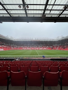 A view from the stands of a large football stadium with an empty field. Rows of red seats extend towards the pitch, which is lush and green. The stadium is open and expansive, with a partially covered ceiling above the spectator area. The words 'Manchester United' are prominently displayed across the seating area on the opposite side.