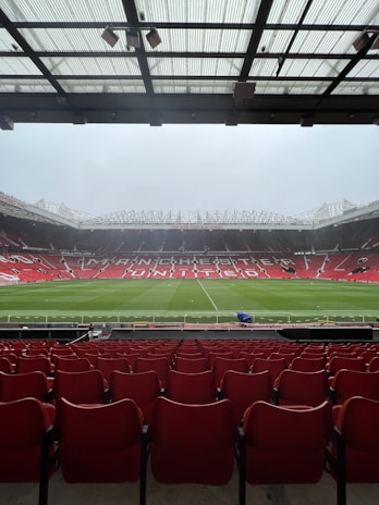 A view from the stands of a large football stadium with an empty field. Rows of red seats extend towards the pitch, which is lush and green. The stadium is open and expansive, with a partially covered ceiling above the spectator area. The words 'Manchester United' are prominently displayed across the seating area on the opposite side.