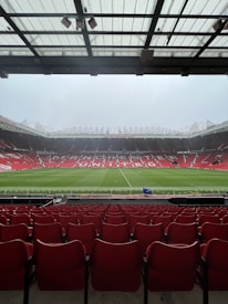 A view from the stands of a large football stadium with an empty field. Rows of red seats extend towards the pitch, which is lush and green. The stadium is open and expansive, with a partially covered ceiling above the spectator area. The words 'Manchester United' are prominently displayed across the seating area on the opposite side.
