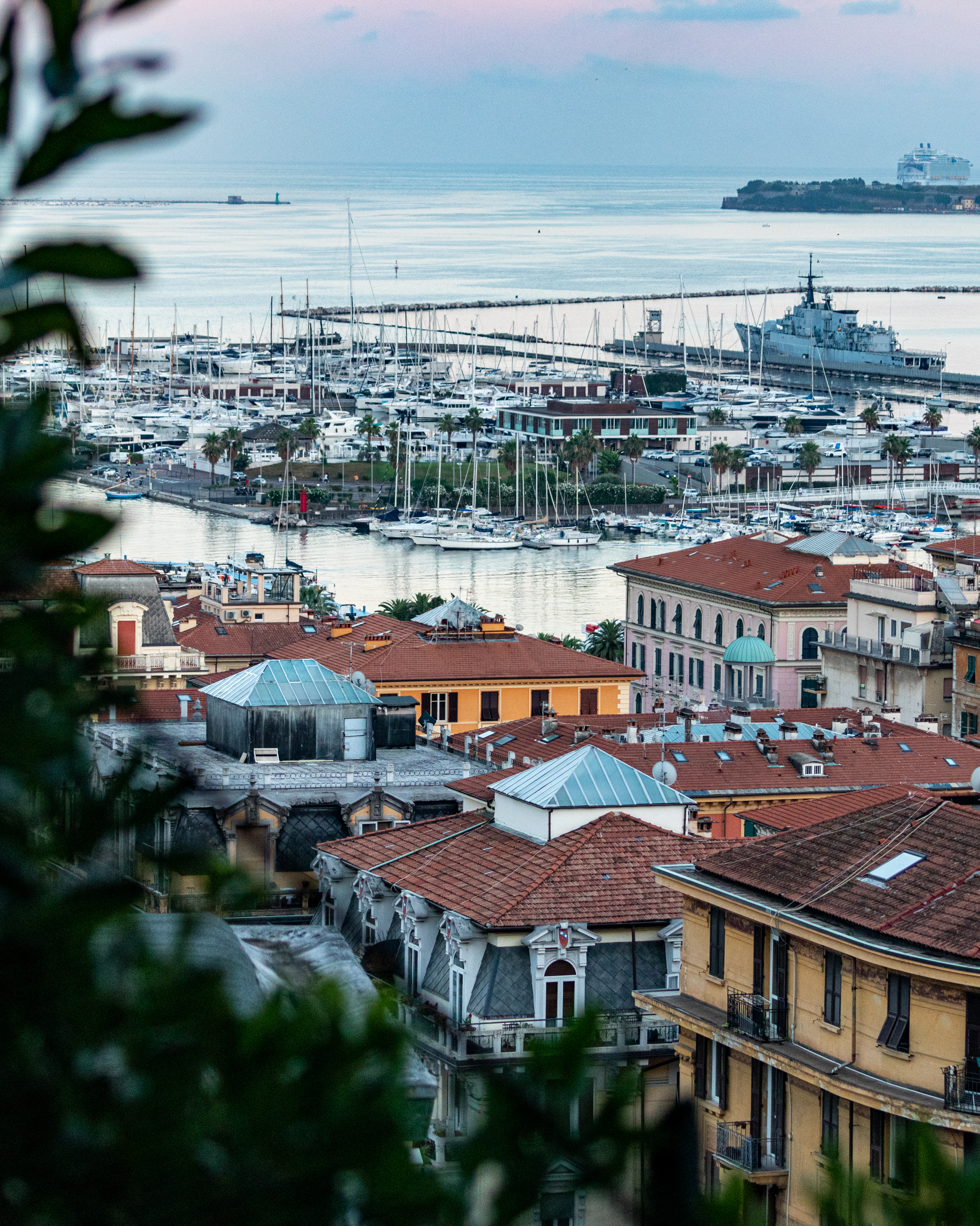 La Spezia at dusk seen over terracotta rooftops: the marina packed with white yachts and sailing masts, a grey naval vessel at the quay and the Gulf of Poets opening out beyond under a soft pink sky