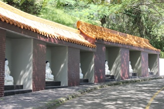 A series of alcoves built into a hillside, each containing a white statue of a seated Buddha. The alcoves have a traditional Asian-style roof with yellow tiles. The surrounding area is lush with greenery, and the sunlight creates patterns of light and shadow on the structures.