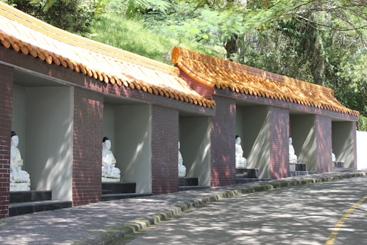 A series of alcoves built into a hillside, each containing a white statue of a seated Buddha. The alcoves have a traditional Asian-style roof with yellow tiles. The surrounding area is lush with greenery, and the sunlight creates patterns of light and shadow on the structures.