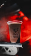 A plastic cup filled with a beverage is placed on a metal surface. The cup has a logo and some text written on it. The background is illuminated with red light, creating a warm and vibrant atmosphere.