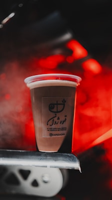 A plastic cup filled with a beverage is placed on a metal surface. The cup has a logo and some text written on it. The background is illuminated with red light, creating a warm and vibrant atmosphere.