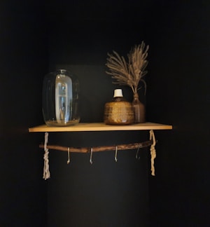 A wooden shelf is mounted on a dark wall, holding decorative items including a large glass jar, a textured ceramic vase with a white top, and a brown vase containing dried plants. Below the shelf, a wooden branch serves as a rod with hooks attached.