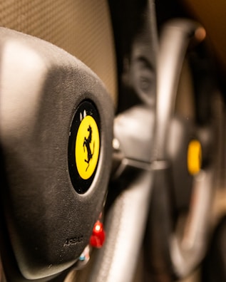 The image focuses on the steering wheel of a luxury sports car, featuring a prominent yellow emblem with a black prancing horse. The leather texture and sleek design elements suggest high-quality craftsmanship.