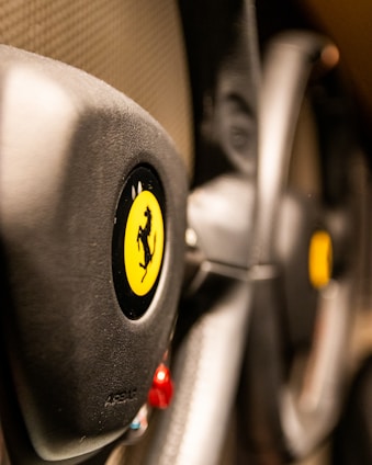 The image focuses on the steering wheel of a luxury sports car, featuring a prominent yellow emblem with a black prancing horse. The leather texture and sleek design elements suggest high-quality craftsmanship.
