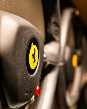 The image focuses on the steering wheel of a luxury sports car, featuring a prominent yellow emblem with a black prancing horse. The leather texture and sleek design elements suggest high-quality craftsmanship.