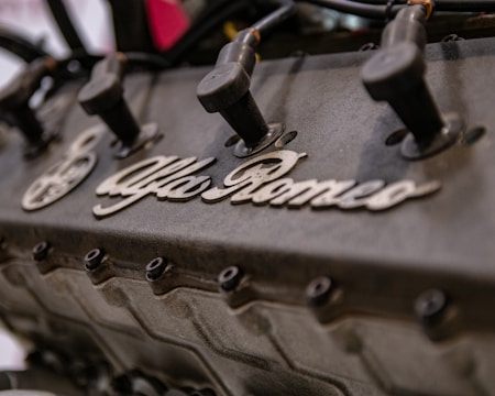 A close-up view of an engine with visible spark plug wires and the Alfa Romeo branding prominently displayed on the engine block.