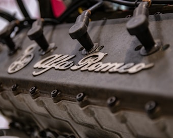 A close-up view of an engine with visible spark plug wires and the Alfa Romeo branding prominently displayed on the engine block.