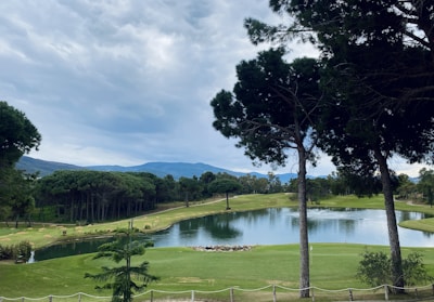 A scenic landscape featuring a well-maintained golf course with lush green grass and a serene pond at the center. Tall trees with dense foliage frame the view, and mountains are visible in the background beneath a cloudy sky.