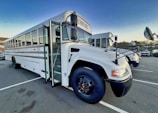 A large, white school bus is parked in an outdoor lot. It has black accents and visible side mirrors. The front door is open, revealing stairs leading into the bus. Another bus is parked behind it.