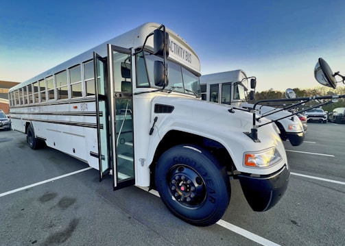 A large, white school bus is parked in an outdoor lot. It has black accents and visible side mirrors. The front door is open, revealing stairs leading into the bus. Another bus is parked behind it.