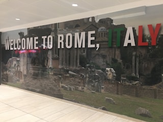 A large, welcoming sign with the phrase 'WELCOME TO ROME, ITALY' in bold letters set against a backdrop of ancient Roman ruins and architecture. The words 'ROME' and 'ITALY' are highlighted with colors of the Italian flag: green, white, and red. The setting is likely an interior space with a tiled floor and overhead lighting.