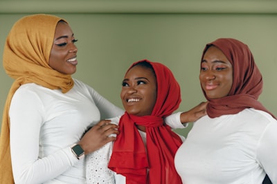 Three people are standing closely together, smiling and interacting with each other. Each is wearing a headscarf—one in mustard, one in red, and one in brown—and white tops. They appear to be enjoying each other's company.