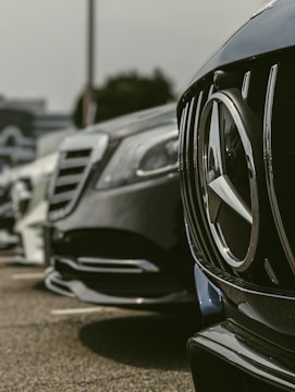 Close-up view of the front ends of several parked luxury cars, highlighting their sleek designs and shiny surfaces. The focus is on the grille and emblem of a black car in the foreground, with other cars lined up in the background.