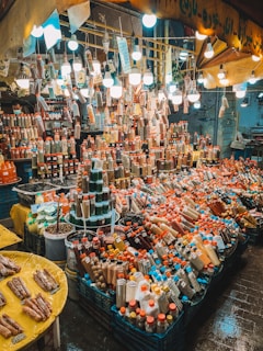 An assortment of spices and dried goods displayed in a vibrant market stall. The scene is illuminated by multiple hanging light bulbs, giving it a warm and lively atmosphere. Numerous bottles and jars filled with colorful spices are neatly organized and cover every surface.