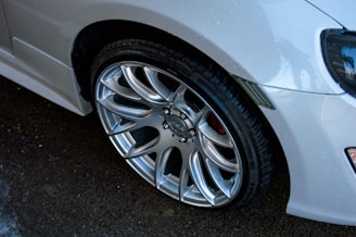 A close-up view of a shiny, silver alloy wheel mounted on a car, with water droplets visible on the car's glossy white body. The image captures the detailed design of the wheel, including its polished surface and intricate patterns.
