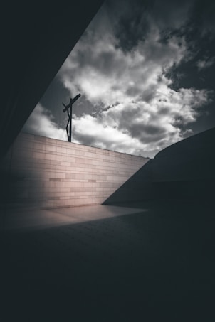 A dramatic scene featuring a cross silhouetted against a cloudy sky. The cross is mounted on a high, light-colored brick wall. The lighting emphasizes the textures and shadows creating a somber atmosphere.