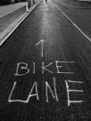 A black and white photograph of a street with the words 'BIKE LANE' and an arrow painted on the asphalt, indicating the designated path for cyclists. The street is flanked by sidewalks and street infrastructure, including a traffic cone and what appears to be a few blurred figures in the distance.