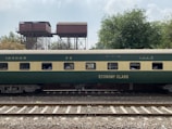 A green and beige passenger train car marked with 'Economy Class' is on railway tracks. In the background, there are old, elevated water tanks and lush trees under a partly cloudy sky. Several passengers can be seen through the windows of the train car.