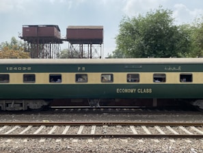 A green and beige passenger train car marked with 'Economy Class' is on railway tracks. In the background, there are old, elevated water tanks and lush trees under a partly cloudy sky. Several passengers can be seen through the windows of the train car.
