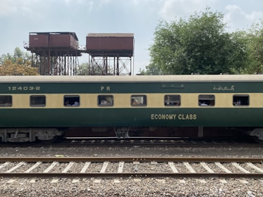 A green and beige passenger train car marked with 'Economy Class' is on railway tracks. In the background, there are old, elevated water tanks and lush trees under a partly cloudy sky. Several passengers can be seen through the windows of the train car.