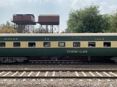 A green and beige passenger train car marked with 'Economy Class' is on railway tracks. In the background, there are old, elevated water tanks and lush trees under a partly cloudy sky. Several passengers can be seen through the windows of the train car.