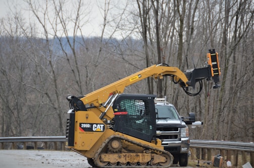 A yellow construction vehicle with a 299D CAT model is parked on a roadside. The vehicle has a large arm extending from its body, equipped with machinery for construction work. In the background, leafless trees and a hilly landscape are visible, indicating a winter or late autumn setting.