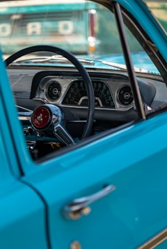 The focus is on the interior view of a vintage car with a turquoise exterior, highlighting the steering wheel, dashboard, and classic speedometer. Visible through the open window is a partial view of another car's emblem reading 'FORD'. The design elements evoke a sense of nostalgia.