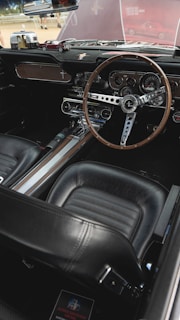 The interior of a vintage car featuring a classic steering wheel with a wooden finish, detailed dashboard with multiple gauges, and a retro design. The seats are upholstered in black leather, and the dashboard includes miniature car models as decorative items.