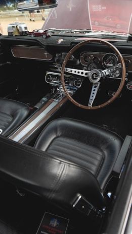 The interior of a vintage car featuring a classic steering wheel with a wooden finish, detailed dashboard with multiple gauges, and a retro design. The seats are upholstered in black leather, and the dashboard includes miniature car models as decorative items.