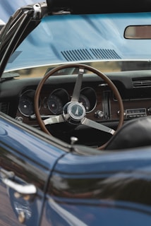 A classic car interior view highlighting a wooden steering wheel, traditional dashboard gauges, and a vintage radio. The blue exterior is visible, suggesting a convertible with the top down.