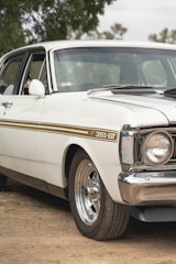 A vintage white car with a gold stripe along its side, marked with 351-GT. The chrome wheel rims and shiny bumper accentuate its classic appeal. It is parked on a dirt surface, surrounded by some greenery in the background.