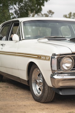 A vintage white car with a gold stripe along its side, marked with 351-GT. The chrome wheel rims and shiny bumper accentuate its classic appeal. It is parked on a dirt surface, surrounded by some greenery in the background.
