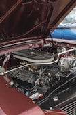 A detailed view of a classic car engine with 'Mustang Powered by Ford' visible on the valve cover. The engine is surrounded by polished metal components and hoses, all set within a burgundy engine bay. The hood is open, revealing the well-maintained mechanics and the overall structure.
