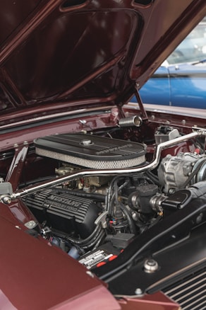 A detailed view of a classic car engine with 'Mustang Powered by Ford' visible on the valve cover. The engine is surrounded by polished metal components and hoses, all set within a burgundy engine bay. The hood is open, revealing the well-maintained mechanics and the overall structure.