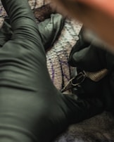 A person wearing black gloves is holding a tattoo machine close to a surface, possibly human skin, with visible tattoo patterns and ink.