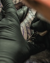 A person wearing black gloves is holding a tattoo machine close to a surface, possibly human skin, with visible tattoo patterns and ink.