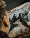 A tattoo artist is in the process of creating a detailed tattoo on a person's back. The artist is wearing black gloves and is holding a tattoo machine wrapped in plastic. There are outlines of intricate designs and patterns visible on the skin, with portions already inked in black.