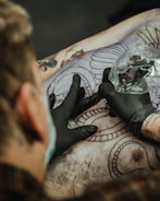 A tattoo artist is in the process of creating a detailed tattoo on a person's back. The artist is wearing black gloves and is holding a tattoo machine wrapped in plastic. There are outlines of intricate designs and patterns visible on the skin, with portions already inked in black.