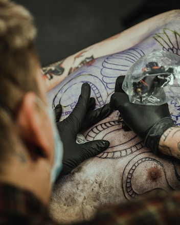 A tattoo artist is in the process of creating a detailed tattoo on a person's back. The artist is wearing black gloves and is holding a tattoo machine wrapped in plastic. There are outlines of intricate designs and patterns visible on the skin, with portions already inked in black.