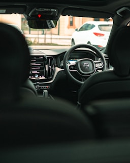 An interior view of a modern car showcasing the steering wheel with a Volvo logo, a digital dashboard, and a central touchscreen display with various controls visible. The car's rearview mirror and window are also in view, revealing a white car parked outside.
