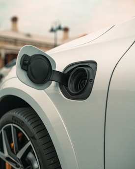A close-up view of an electric car's charging port with the cover open, displaying the intricate design and details of the port. The car is white, and a Pirelli tire is visible, indicating a focus on the wheel area. The background includes a blurred building and sky, suggesting an outdoor setting.